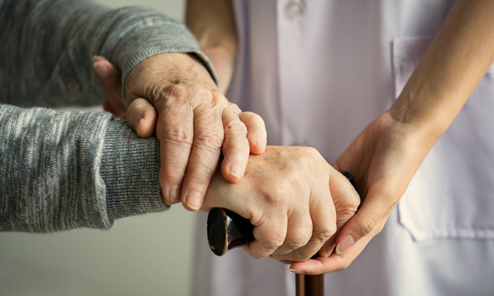 Frail old hands on a walking stick being supported by a nurse